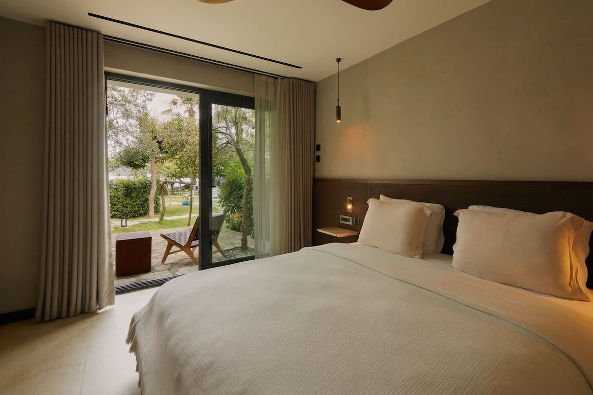 Habitación de hotel con una cama grande, un ventilador de techo, una mesita de noche, una lámpara y vistas al exterior.