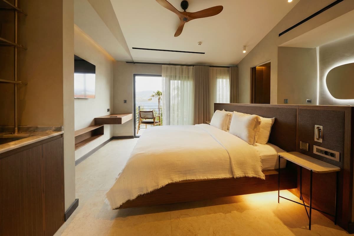 Una habitación de hotel moderna con una cama grande, un ventilador de techo, un pequeño balcón con una silla y un espejo amplio.