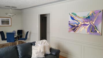 A living room with a black leather sofa, a glass dining table with blue chairs, and a colorful abstract painting on the wall.
