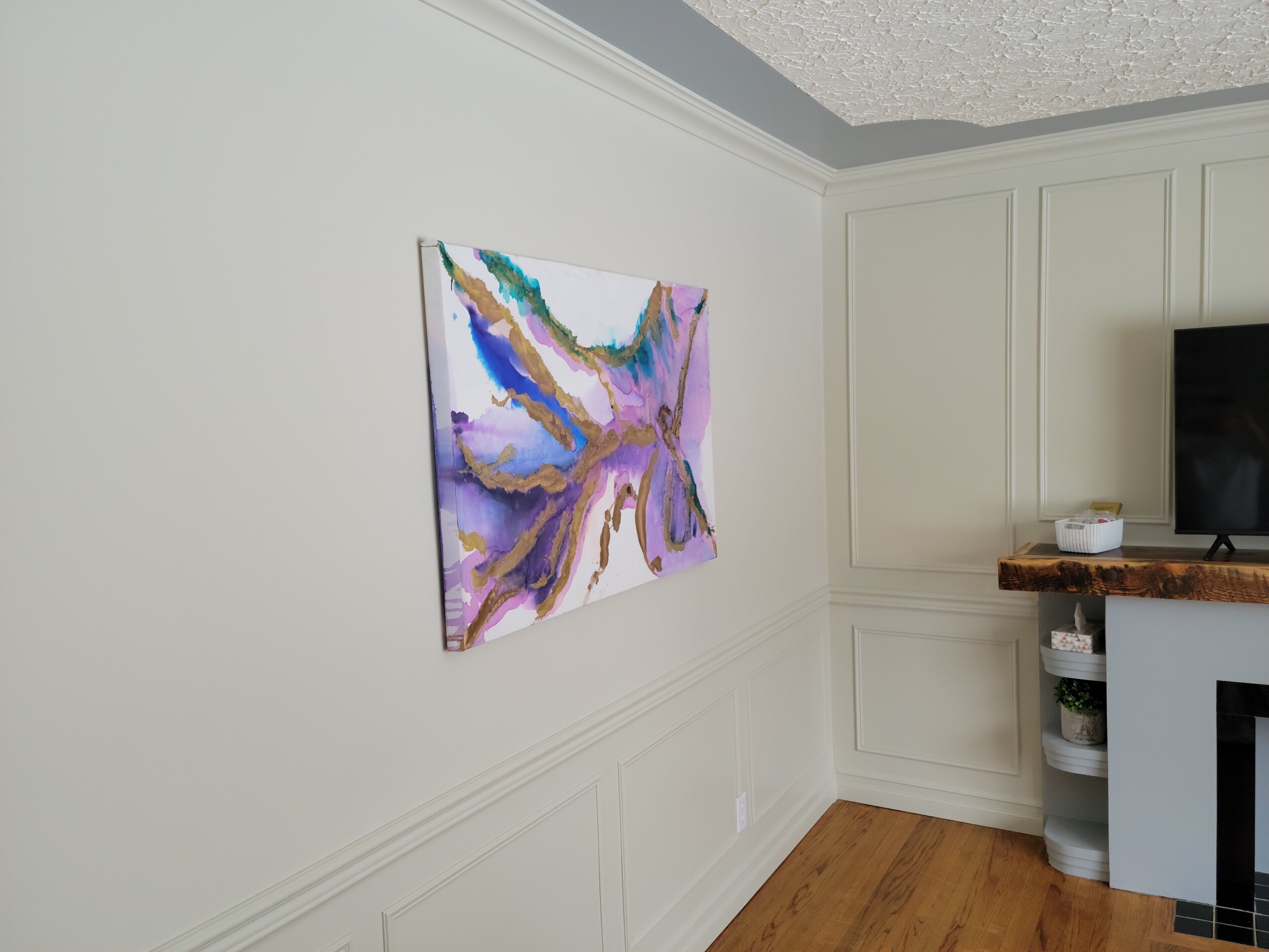 Une pièce avec un mur blanc, un tableau abstrait accroché au mur, un plancher en bois et un meuble blanc contenant un téléviseur.