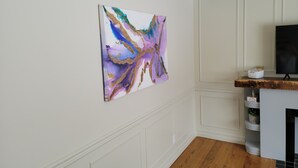Ein Zimmer mit weißer Wand, einem aufgehängten abstrakten Gemälde, Holzboden und einem weißen Schrank mit Fernseher.