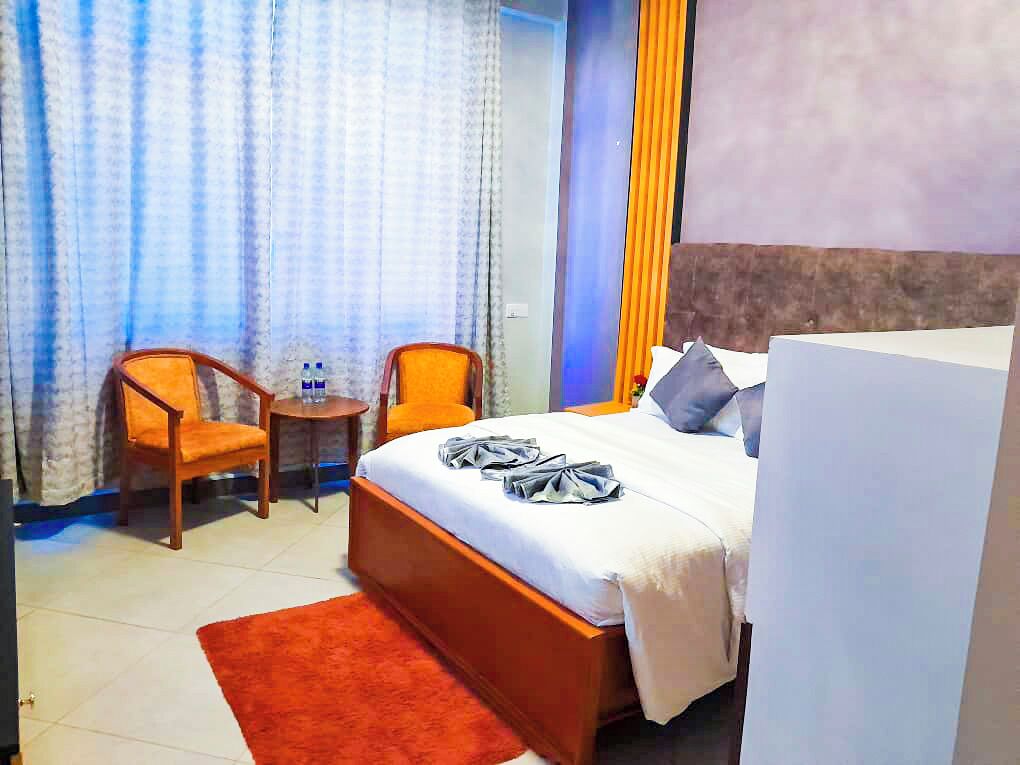 Habitación de hotel con cama, dos sillas, una mesita, alfombra roja, cortinas y una pared con rayas verticales.
