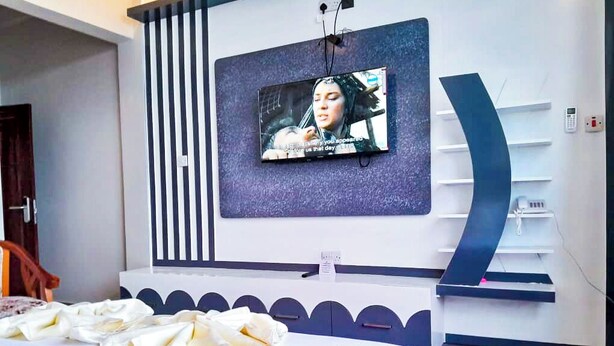A modern hotel room with a large bed, a flat-screen TV mounted on the wall, and a built-in shelving unit. - RIVANA HOTEL GOBA (Dar es Salaam)