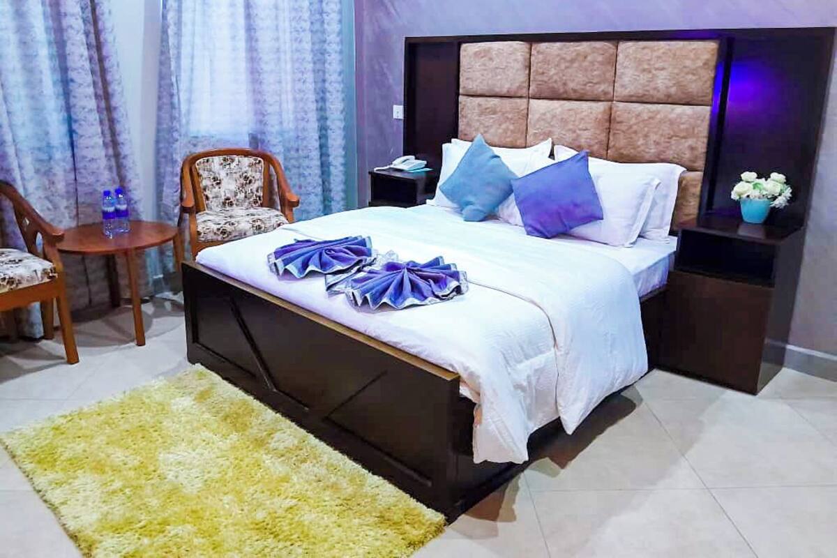 A neatly made bed with a headboard, a bedside table with a lamp, a chair, and a small table with a vase of flowers.