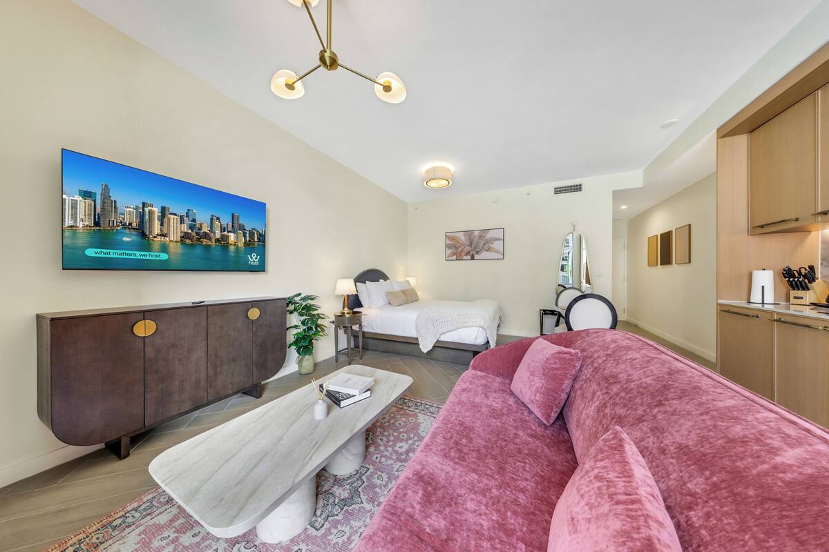 Una sala moderna con una televisión mostrando un paisaje urbano, una cama, un sofá rosa, un mueble de madera y una zona de comedor.