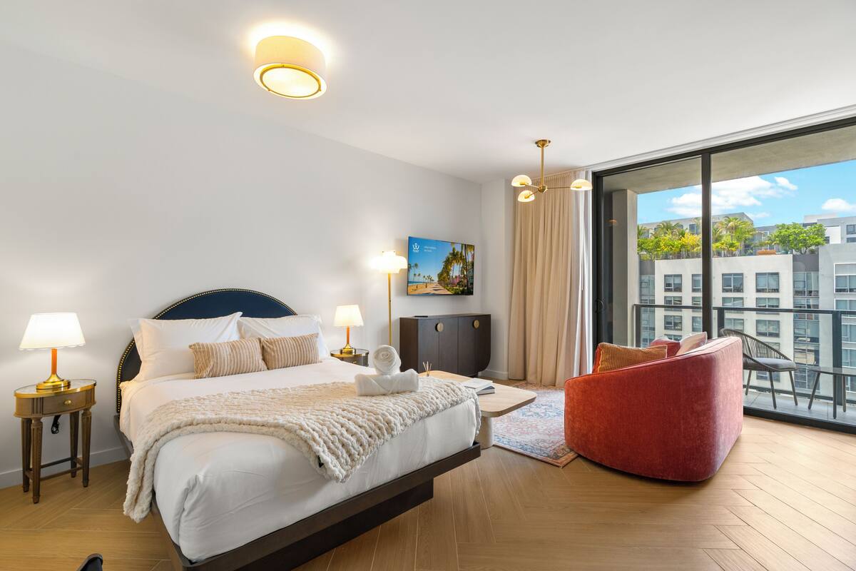 Una habitación de hotel moderna con una cama grande, un sillón rojo, vistas a la ciudad y un balcón con barandilla.