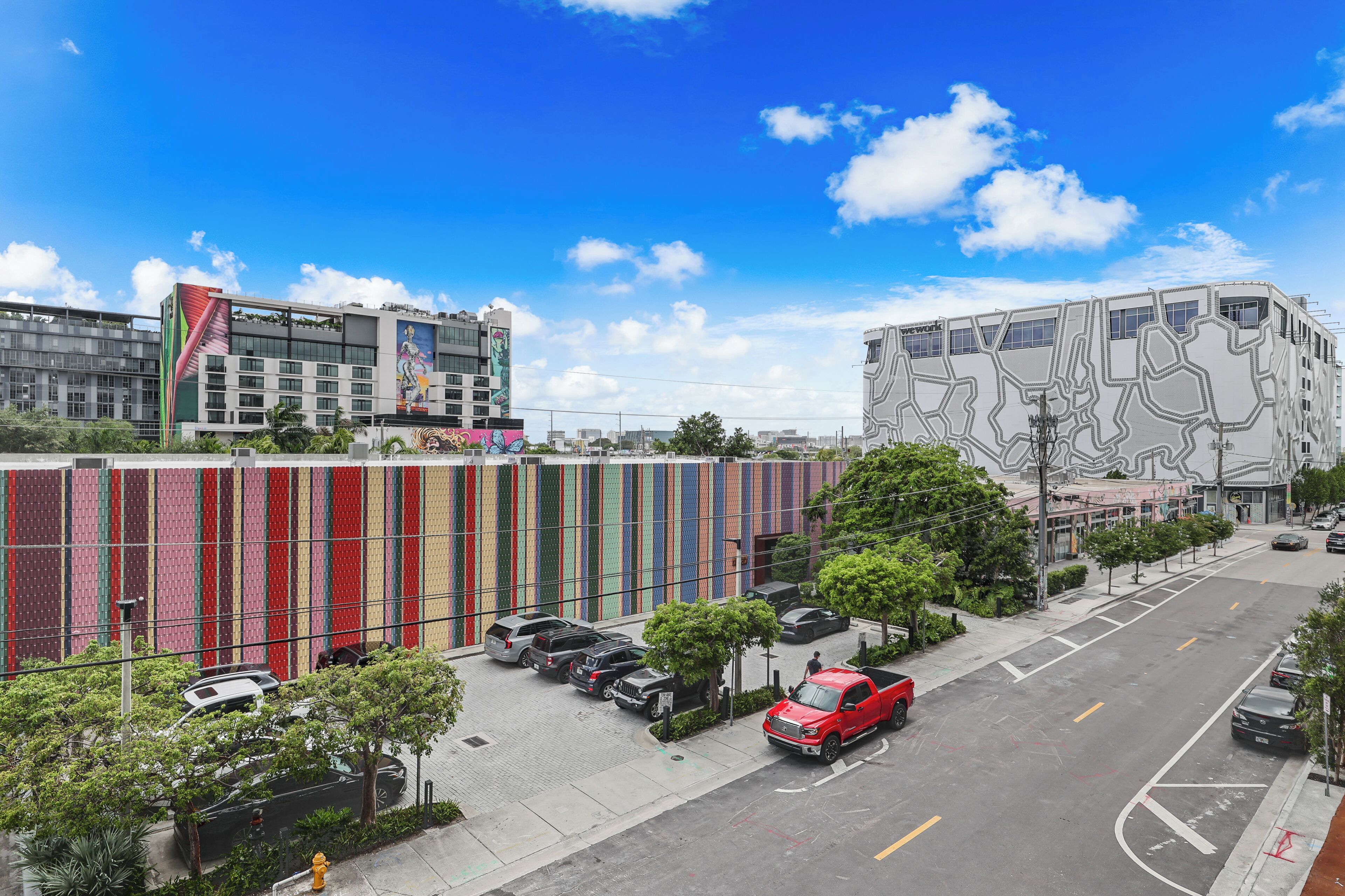 Un immeuble à plusieurs étages avec une façade aux couleurs vives, un parking où sont garées plusieurs voitures et un ciel d’un bleu éclatant.