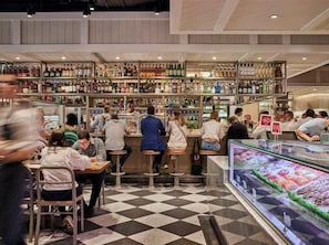 Un espace bar doté de plusieurs étagères remplies de bouteilles, de tabourets de bar et d’une vitrine où sont présentés des produits alimentaires.