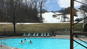Ein Schwimmbad mit darin badenden Personen, umgeben von einer Ziegelterrasse, und eine Skipiste im Hintergrund.