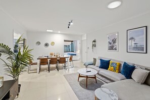 Una sala moderna con un sofá, una mesa de centro, una mesa de comedor y sillas. Hay un ventanal con cortinas, una planta y decoración en la pared.