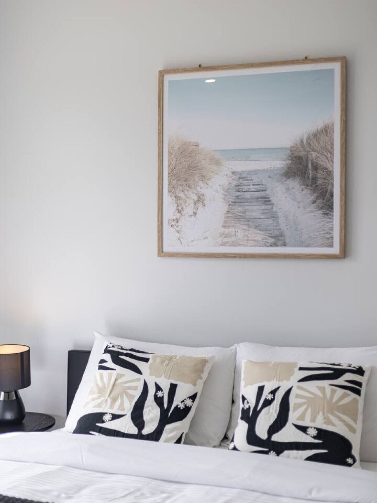 Een slaapkamer met een bed, twee kussens met een natuurprint, een ingelijst beeld van een strandpad en een bedlamp.
