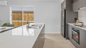 A modern kitchen with a large island, stainless steel appliances, and a view of a wooden fence outside. - Brand New Family Home | Space, Comfort & Location (Park Ridge)