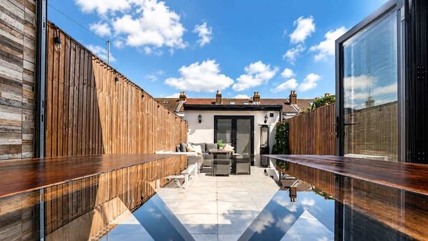 A modern outdoor patio with a wooden fence, a glass balustrade, and a view of a house in the background. - Charming Portsmouth Home w/ Private Garden (Portsmouth)
