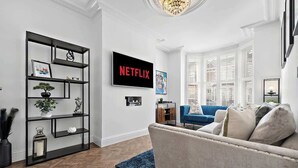 A modern living room with a blue sofa, a black bookshelf, a TV mounted on the wall, and a window with blinds. - Charming Portsmouth Home w/ Private Garden (Portsmouth)