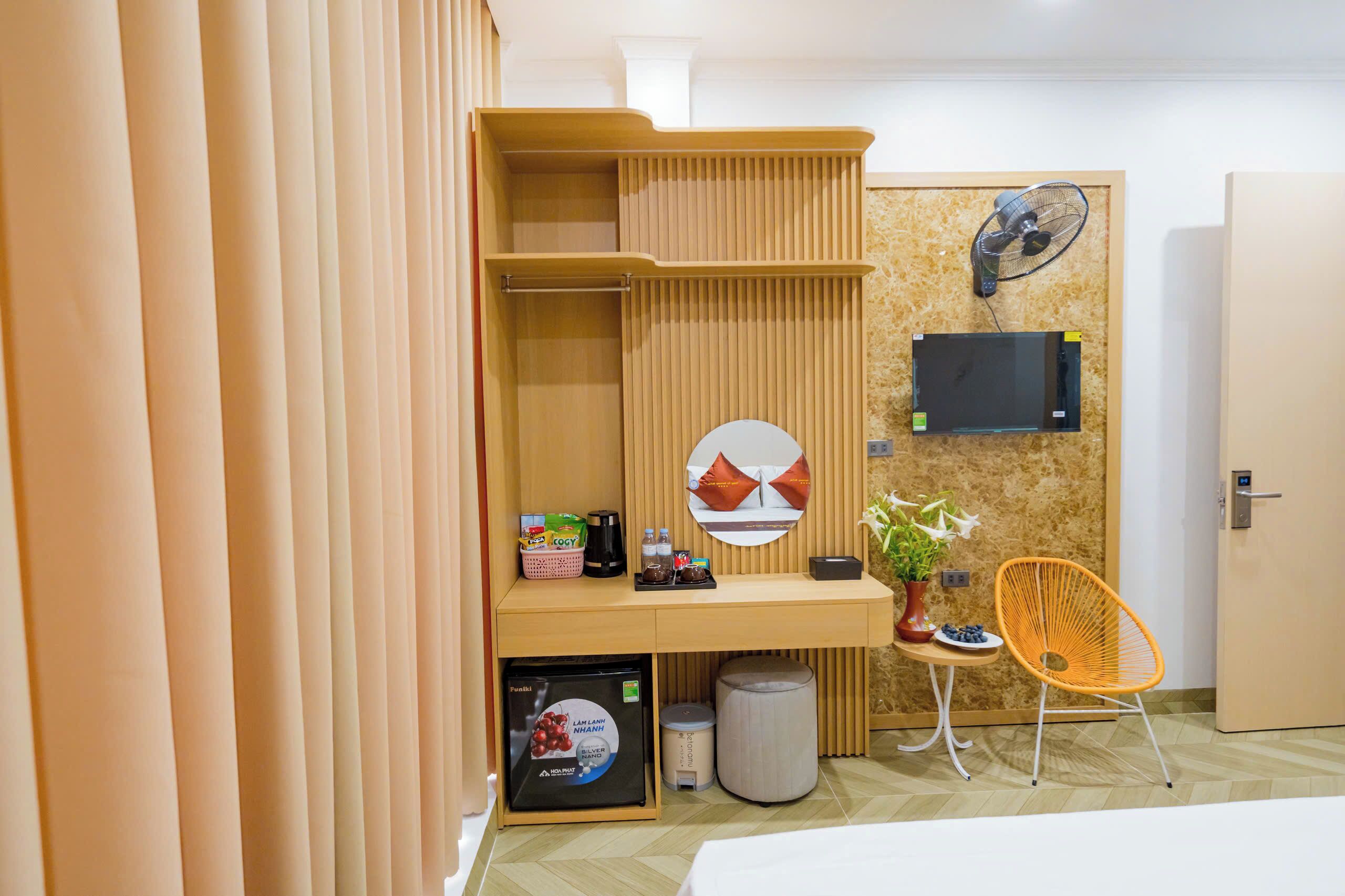A compact hotel room with a built-in desk, a small refrigerator, a microwave, a fan, a potted plant, and a chair.
