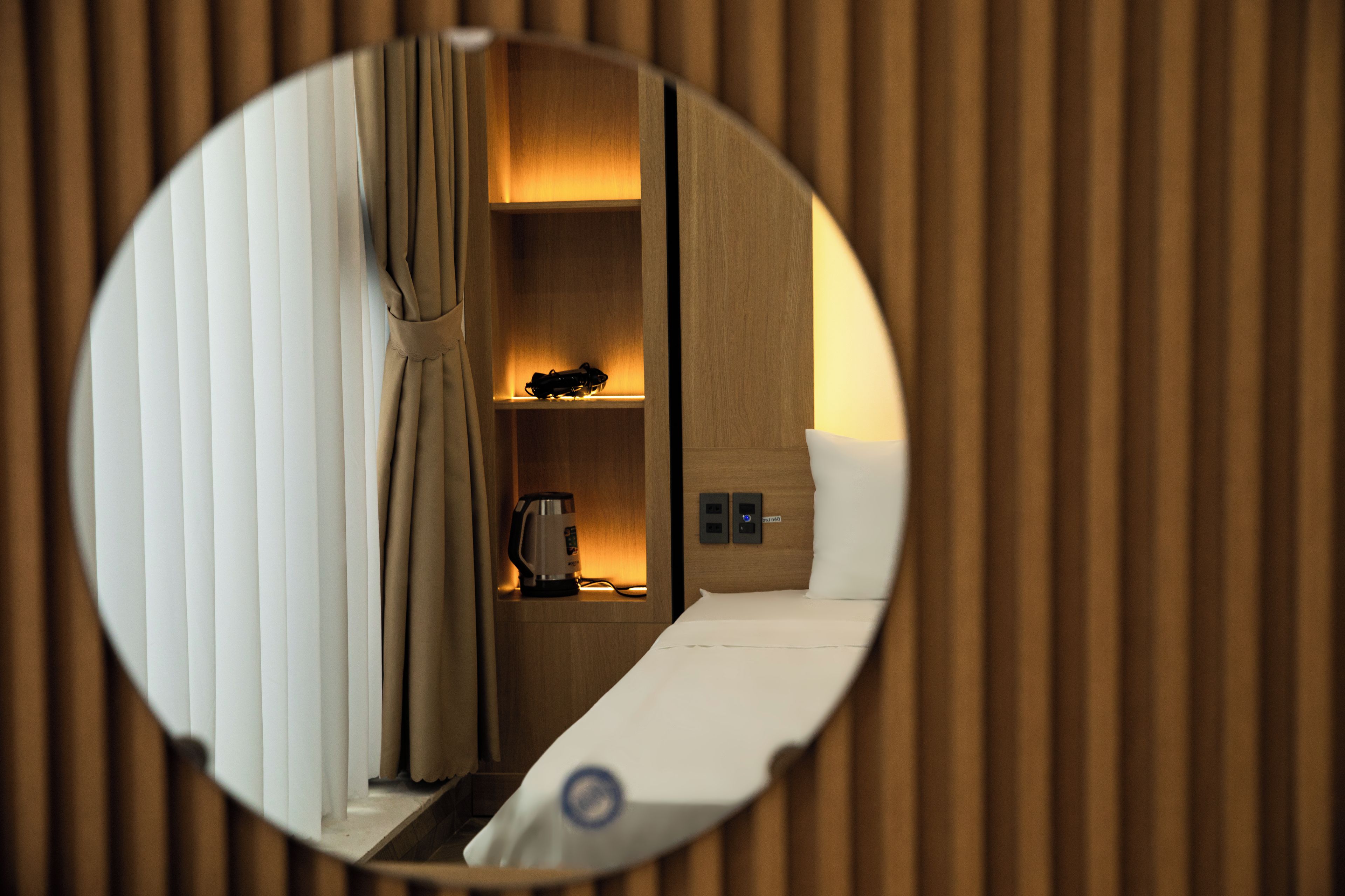 A circular mirror reflecting a room with a bed, a nightstand, a kettle, and a power outlet.