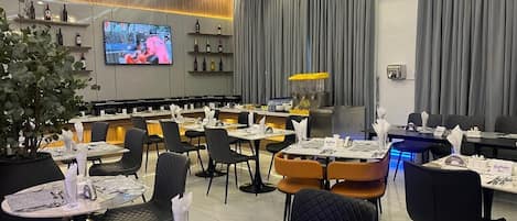 A restaurant with round tables, black chairs, a TV screen, and a bar area with shelves of bottles.