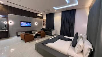 A modern hotel room with a large bed, a seating area with a sofa and armchairs, a flat-screen TV, and a kitchenette.