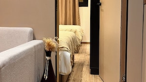 Um corredor estreito com uma cama, uma mesa de cabeceira com um vaso, e uma porta que dá para outro quarto.