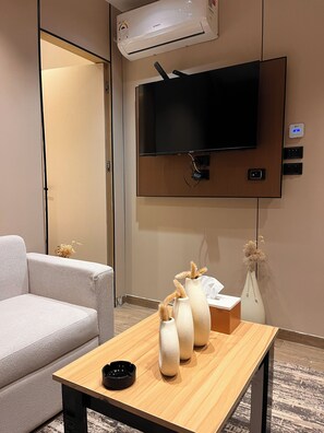 A room with a flat-screen TV mounted on the wall, a wooden table with three vases, a sofa, and an air conditioning unit. - Serene studio with AC/Netflix in wonderful 6 October city (6 October)