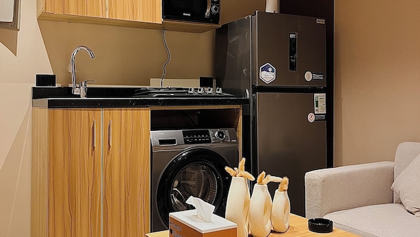Uma cozinha compacta com micro-ondas, máquina de lavar e frigorífico. Há uma pequena mesa com uma caixa com a inscrição 'Home Sweet Home' e objetos decorativos.