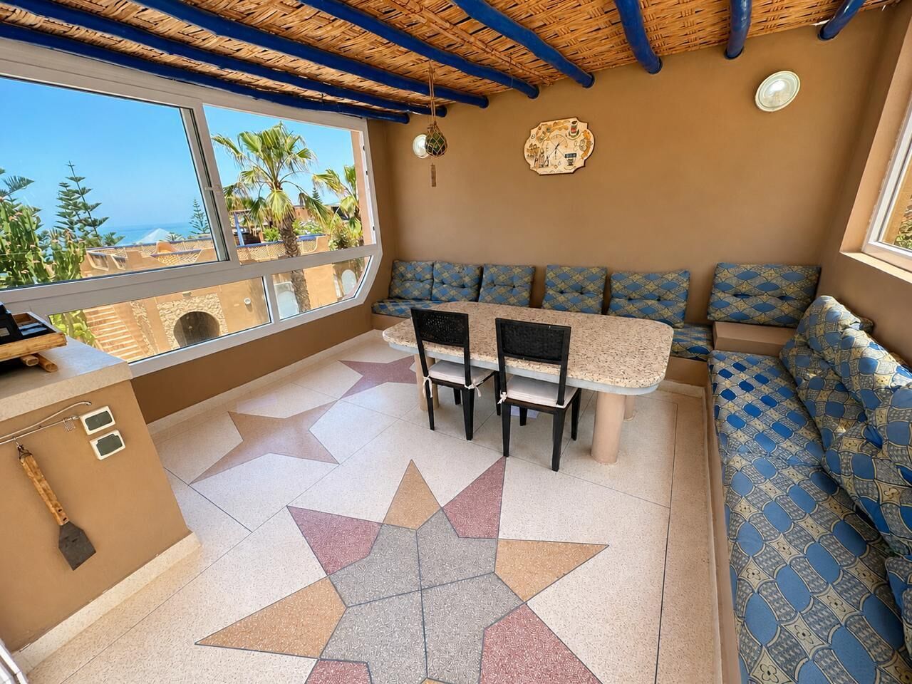 Ein Zimmer mit einem sternenmusterbelagten Boden, einem Tisch mit Stühlen, einer Couch, einem Fenster mit Blick auf Palmen und das Meer sowie einer Wanduhr.