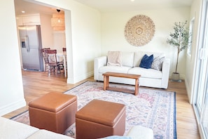 A living room with a sofa, ottomans, a coffee table, a patterned rug, a decorative wall piece, and a dining area in the background.