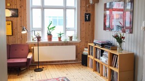 Una habitación con piso de madera, una ventana con persianas, un radiador, una estantería, un sillón morado y una lámpara de pie.