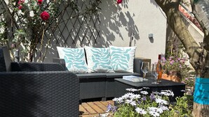 Et udendørs siddeområde med fletmøbler, et bord, hynder og en potteplante.