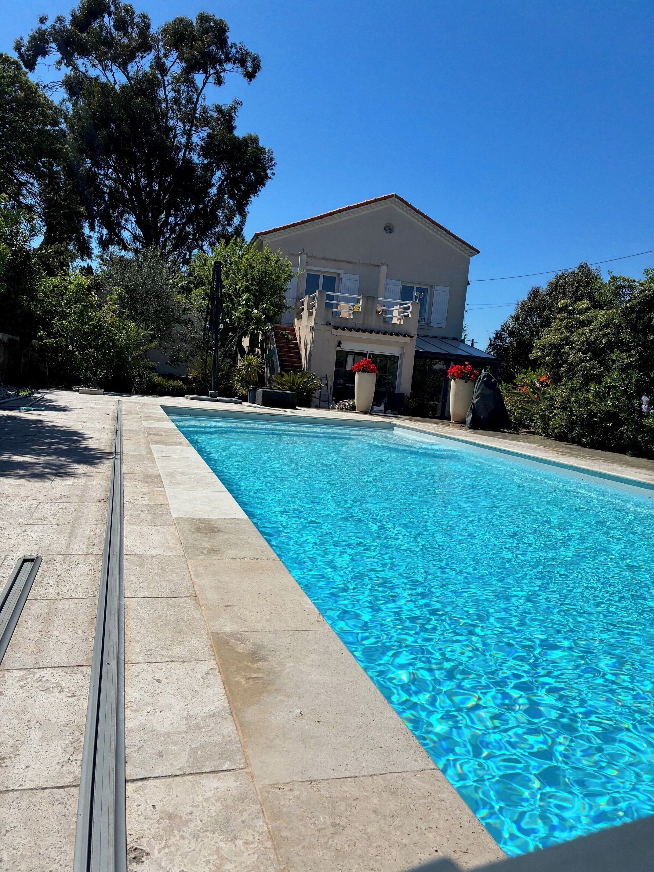 A swimming pool with a clear blue water surface, surrounded by a stone deck. In the background, there is a two-story building with a balcony.