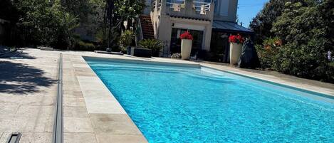 A swimming pool with a clear blue water surface, surrounded by a stone deck. In the background, there is a two-story building with a balcony.