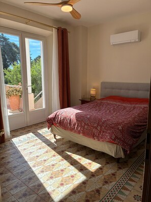 Une chambre à coucher comprenant un lit, un ventilateur de plafond, un climatiseur, une table de chevet et une vue sur l’extérieur, accessible par une porte-fenêtre coulissante.