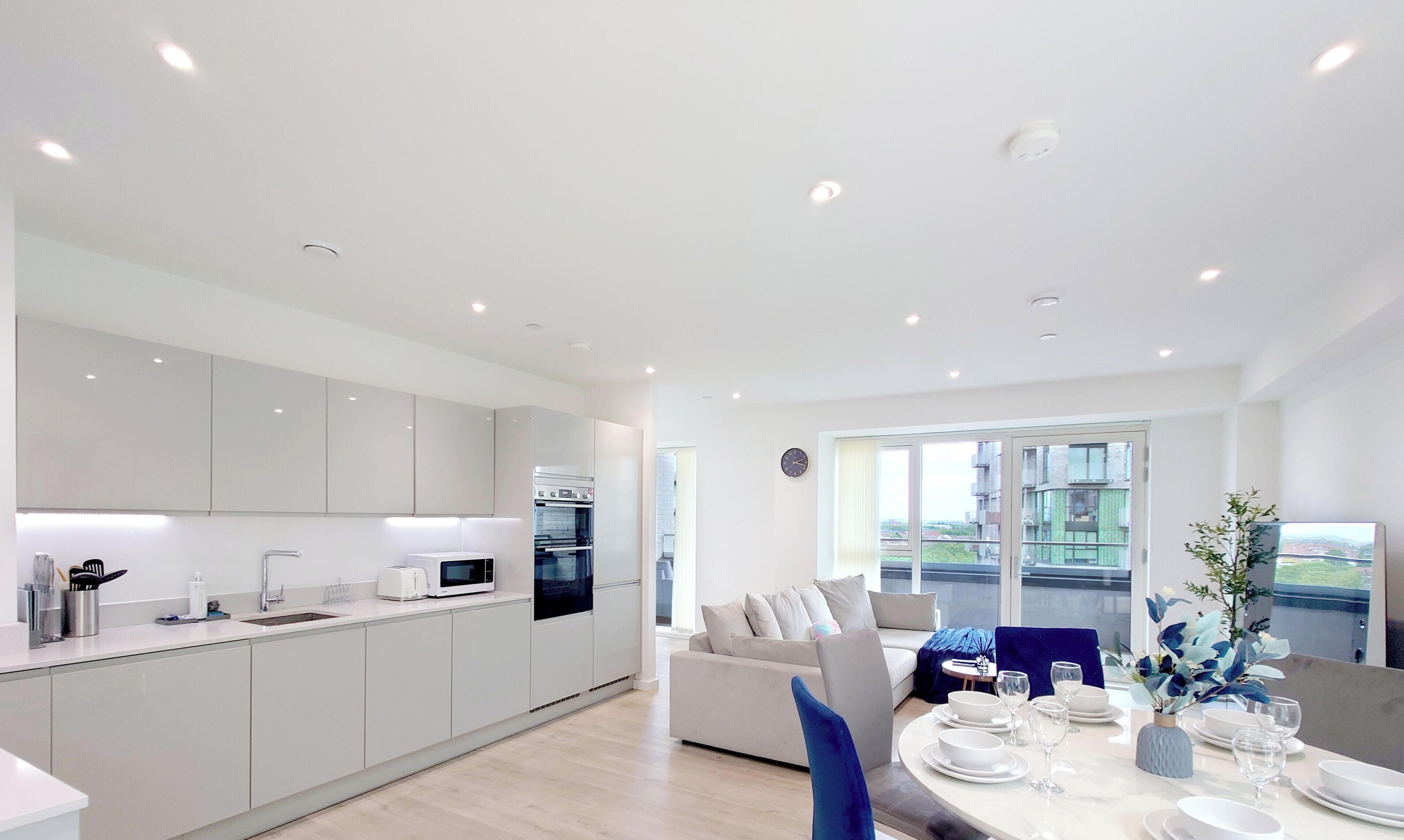 A modern kitchen with white cabinets, a dining area with a table set for four, and a view of buildings through large windows.
