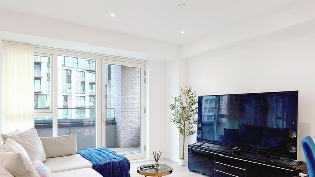 A modern living room with a sofa, a flat-screen TV, and a large window offering a view of a building. - Deluxe Serviced Three Bedroom Apartment (London)