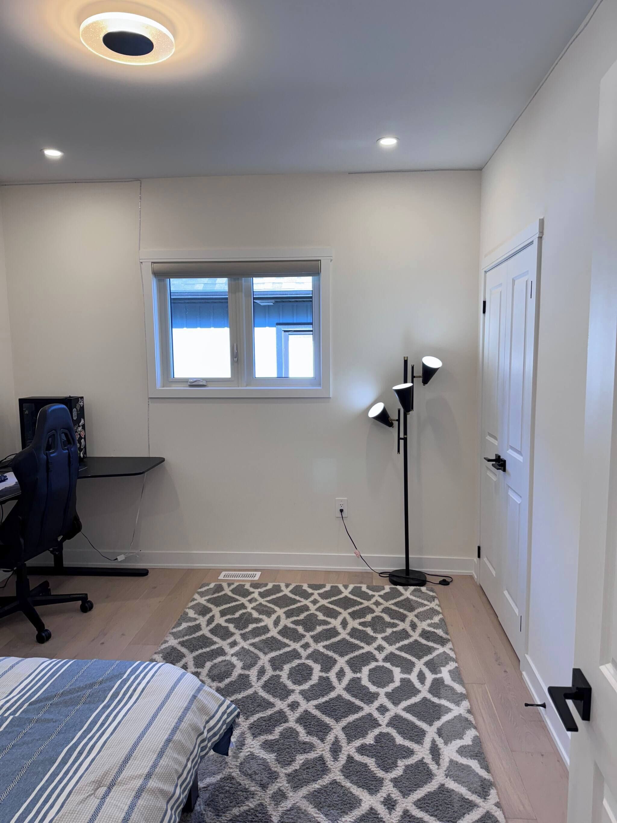 A bedroom with a bed, a desk, a chair, a standing lamp, a window, and a door.