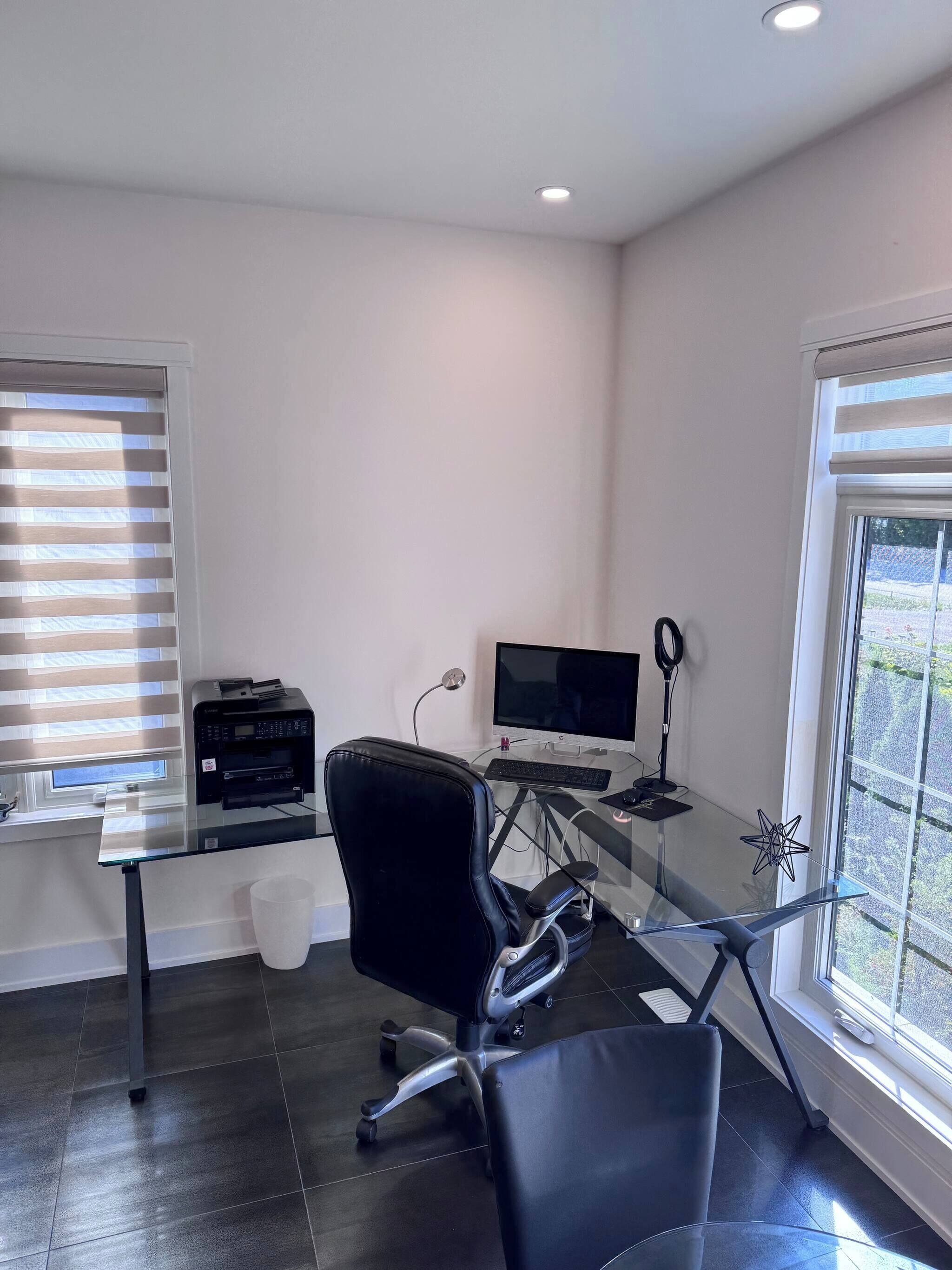 A modern office with a glass desk, black office chair, printer, computer, and large windows with blinds.