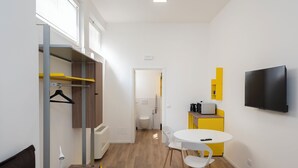 Una habitación de hotel moderna y funcional con vestidor, un pequeño comedor con mesa y sillas, televisor montado en la pared y un baño visible con inodoro y ducha.