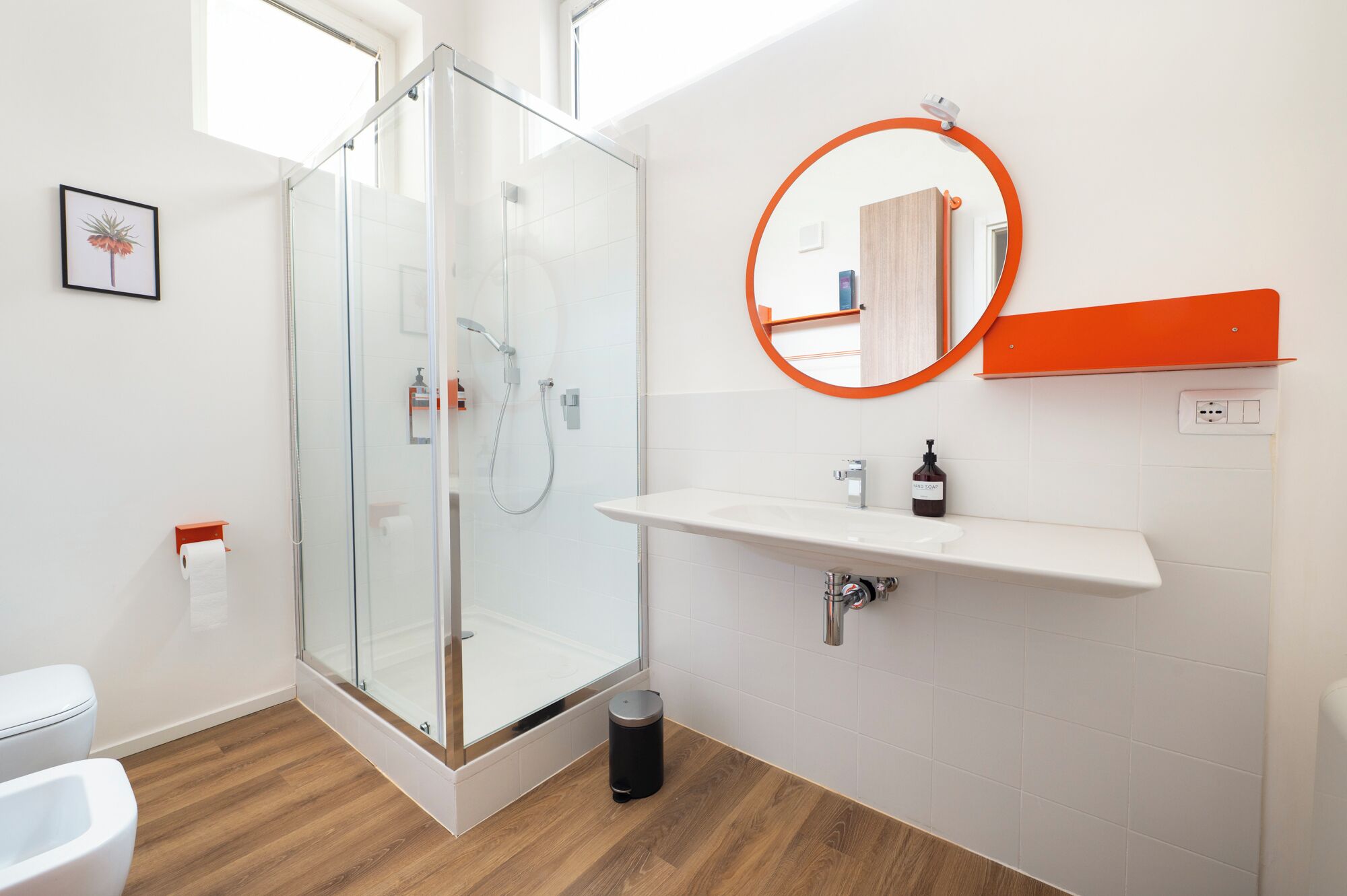 Un baño moderno con cabina de ducha de vidrio, un lavabo blanco con dispensador de jabón y un espejo redondo con marco naranja.