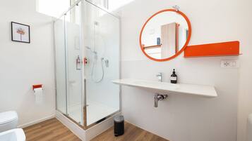 Un baño moderno con cabina de ducha de vidrio, un lavabo blanco con dispensador de jabón y un espejo redondo con marco naranja.