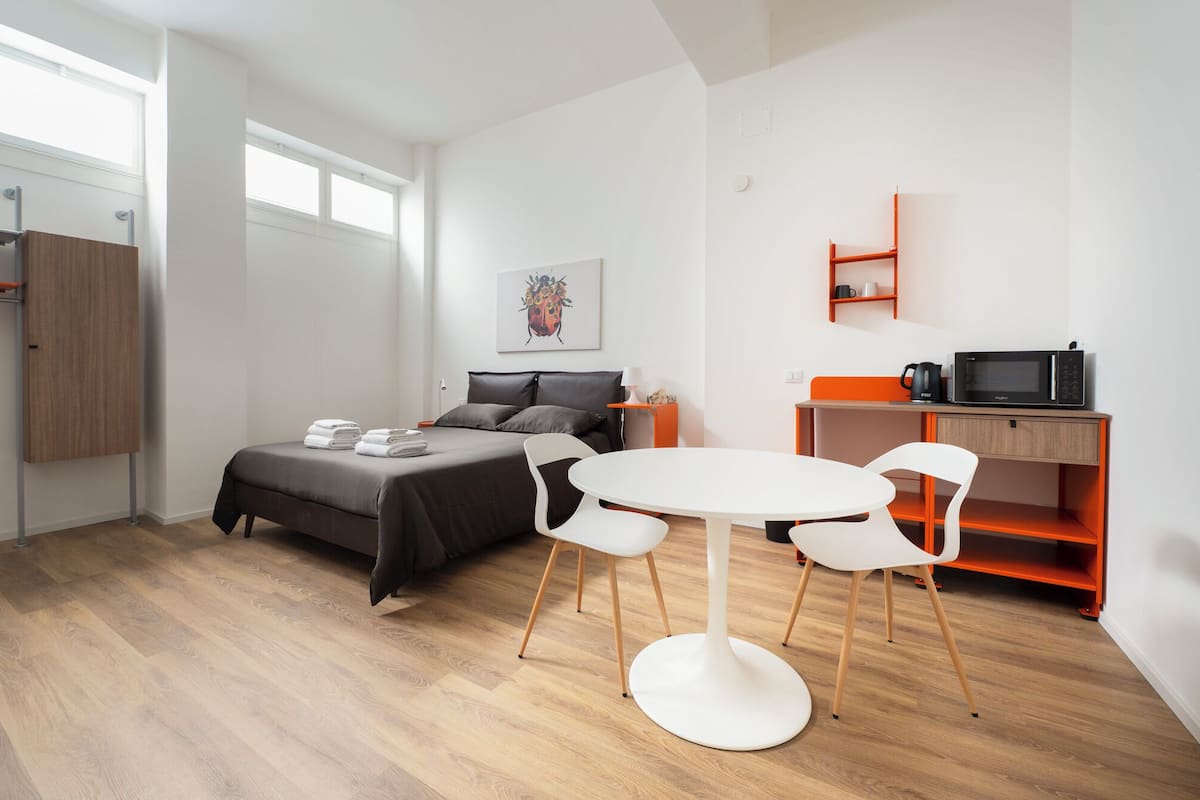A modern, well-lit room with a bed, a round table with chairs, a small desk with a microwave, and a cabinet.