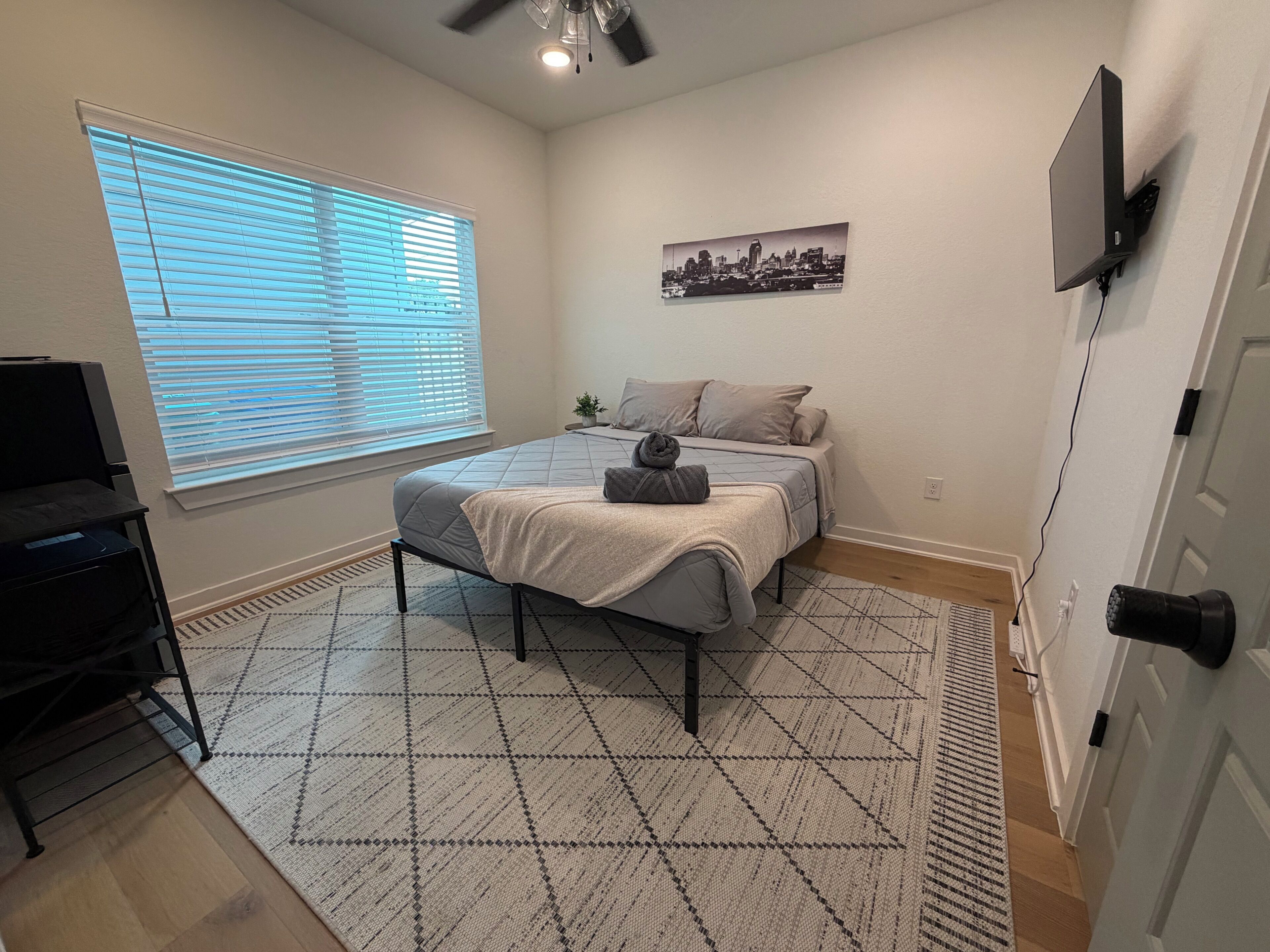 Une chambre à coucher comprenant un lit, un bureau, une chaise, un ventilateur de plafond, une fenêtre avec des stores, un téléviseur fixé au mur et un tableau représentant un paysage urbain.