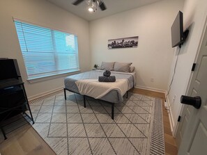 Ein Schlafzimmer mit Bett, Schreibtisch, Stuhl, Deckenventilator, Fenster mit Jalousien, einem an der Wand montierten Fernseher und einem Stadtbild.