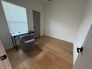 Ein Zimmer mit Holzboden, Schreibtisch mit Stuhl, Fenster mit Jalousien und einer Tür.