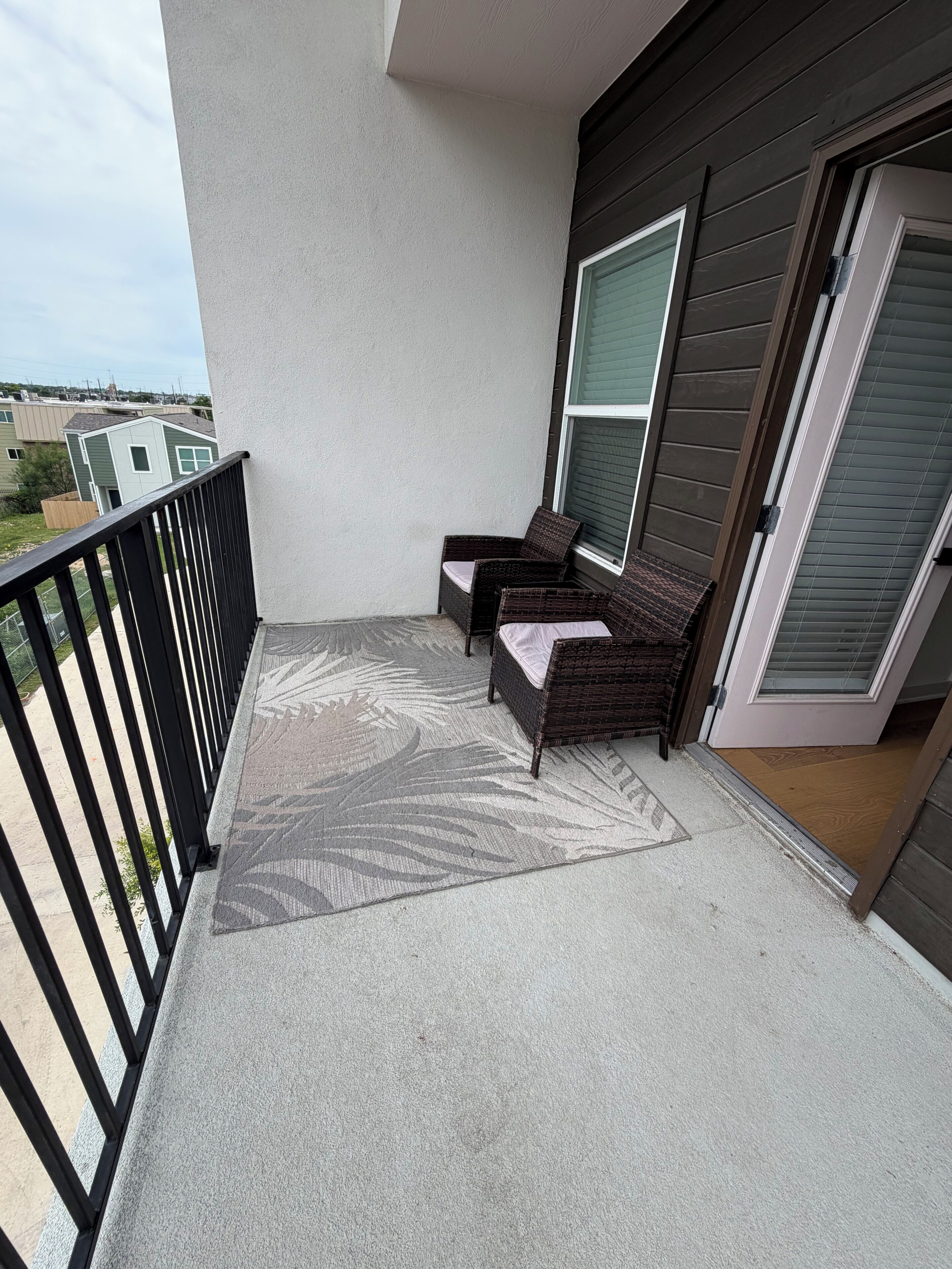 A balcony with two wicker chairs, a patterned rug, and a view of neighboring houses.