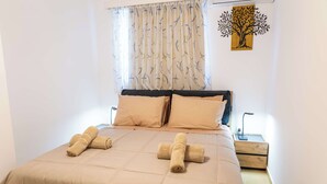 Un lit bien fait, avec une literie beige, une table de chevet en bois sur laquelle se trouve une lampe, et une œuvre d’art représentant un arbre, accrochée au mur.