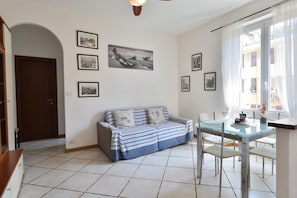 A living room with a sofa, a dining table with chairs, framed pictures on the wall, and a window with curtains.
