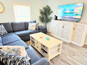 Ein Wohnzimmer mit einem Ecksofa, einem Fernseher, einem Schrank und einer Pflanze.