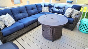 Un canapé d’angle avec une table ronde au centre, un fauteuil en osier et une petite poubelle sur une terrasse en bois.