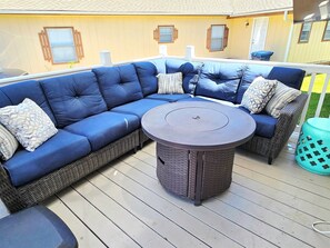 Ein Ecksofa mit einem runden Tisch in der Mitte, ein Korbstuhl und ein kleiner Mülleimer auf einer Holzterrasse.