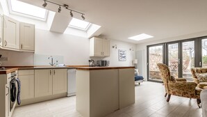 A modern kitchen with a central island, a dining area with floral chairs, and a large sliding glass door leading to an outdoor space. - Devon Cottage | Elie (Elie)